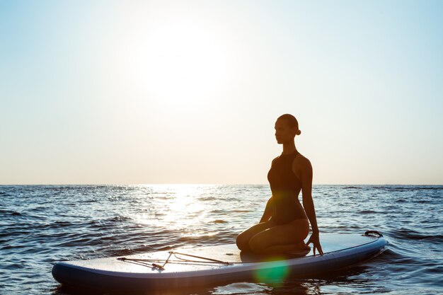 Die Silhouette einer sch&ouml;nen Frau macht Yoga auf einem Surfbrett bei Sonnenaufgang 176420 2034